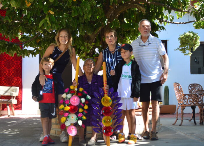 Six people of different ages pose together outdoors under a leafy tree, with two colorful floral arrangements in front of them—capturing the joy of an authentic food tour. A white building and courtyard furniture complete the inviting scene.