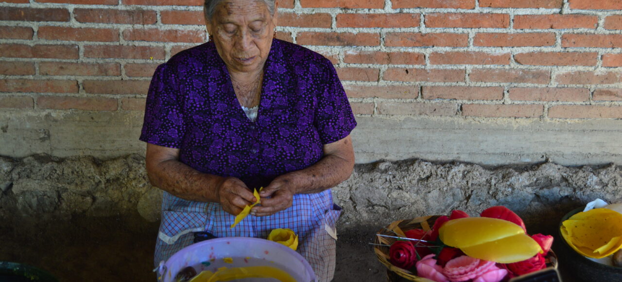 An elderly woman in a purple blouse shapes yellow wax by hand, sitting by a brick wall with wax figures and flowers—offering a glimpse into the artistry celebrated on Mexico Foodie Tours for an authentic culinary experience.