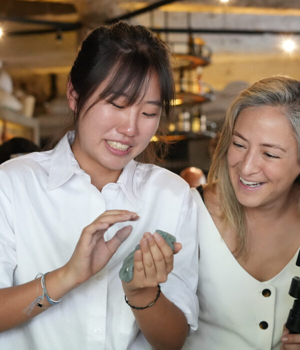 Two women stand indoors; one holds a phone and gestures while the other smiles and holds a tripod, ready to capture their Mexico Foodie Tours culinary experience. Shelves and people are visible in the blurred background. Two women stand indoors; one holds a phone and gestures while the other smiles and holds a tripod, ready to capture their Mexico Foodie Tours culinary experience. Shelves and people are visible in the blurred background.
